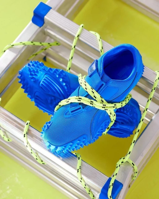Pair of bright blue running shoes tied together with neon yellow cord, resting on an aluminum step ladder against a bright green backdrop.