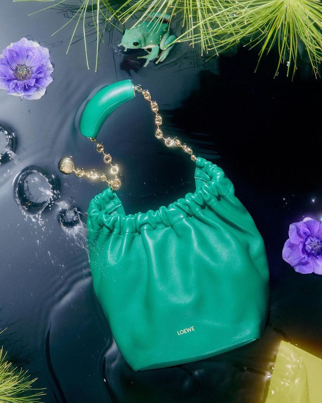 A green Loewe handbag floats in water with scattered purple flowers, reeds, and a decorative frog.
