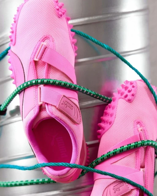 Close-up of pink Puma sneakers with a textured sole and green elastic cord, showcasing a fashion and sports style against a metallic background.