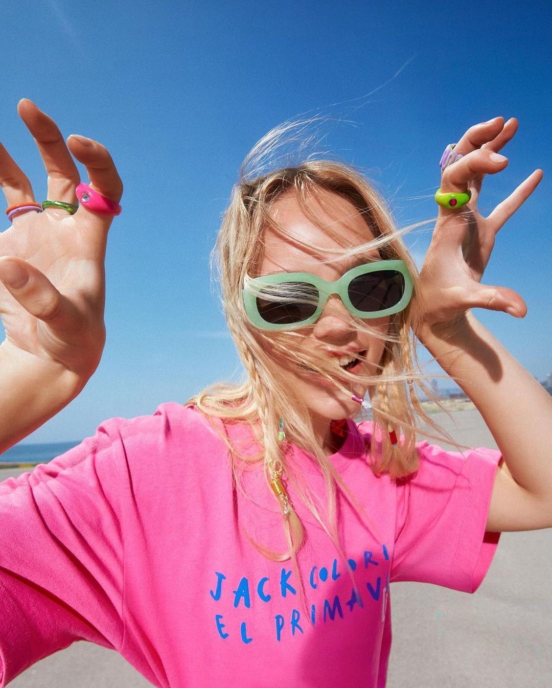 A woman smiles and holds her hands up wearing colorful rings and sunglasses in a sunny outdoor setting, posing in front of a blue sky.