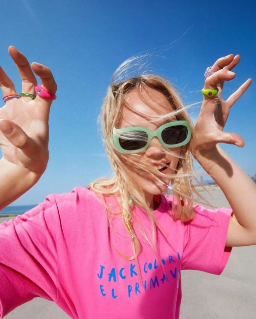 A woman smiles and holds her hands up wearing colorful rings and sunglasses in a sunny outdoor setting, posing in front of a blue sky.
