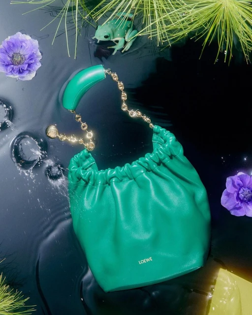Green leather handbag with gold chain on reflective wet surface, adorned with flowers and a decorative frog figurine.