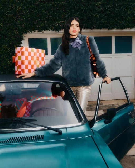 Adult female poses next to a teal car filled with gifts, garage doors and ivy in the background, fashion and automotive photography.
