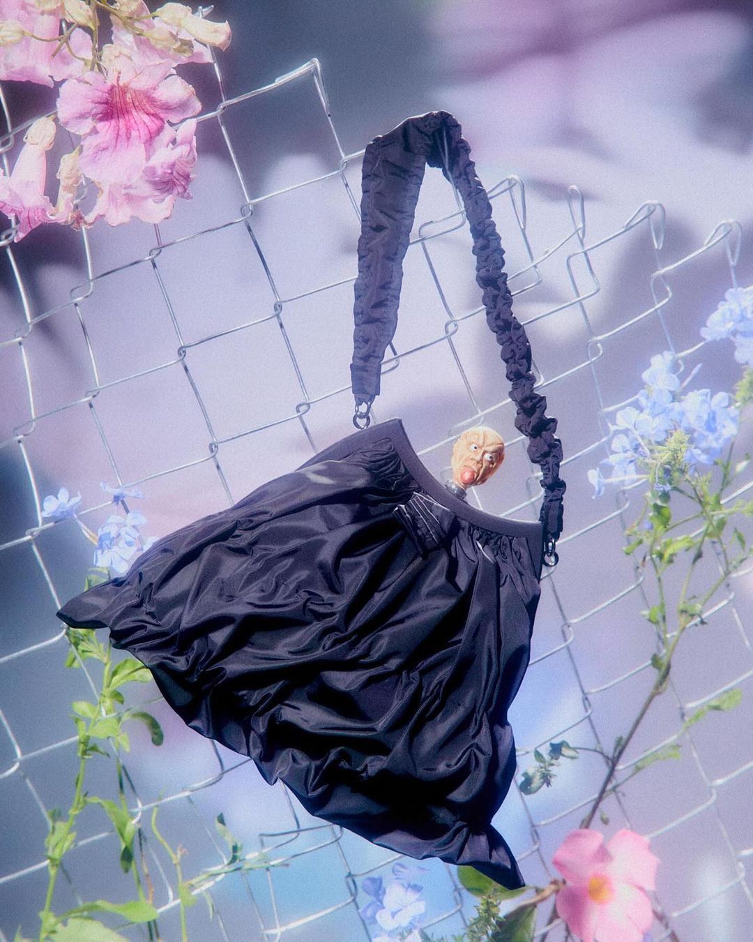 Elegant black purse with gathered details hangs against a wire fence, surrounded by pink and blue flowers, creating a surreal fashion shot.