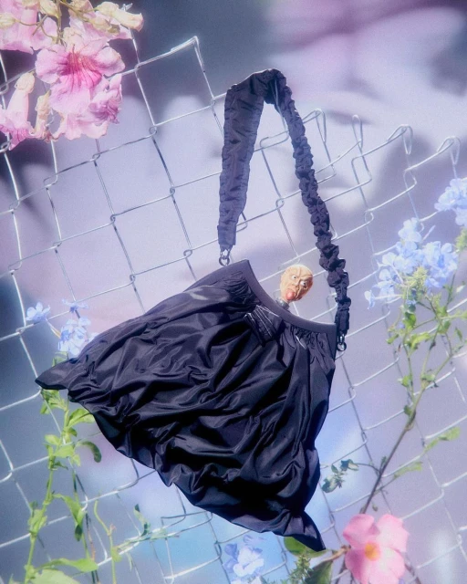 Elegant black purse with gathered details hangs against a wire fence, surrounded by pink and blue flowers, creating a surreal fashion shot.