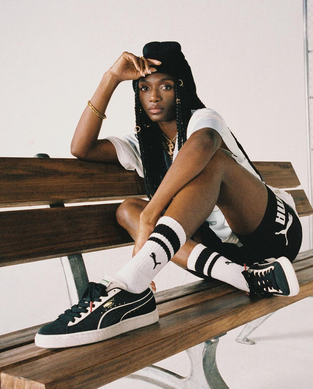 Athletic woman sits on bench wearing Puma sneakers and shorts with logo socks; neutral studio backdrop and soft lighting.