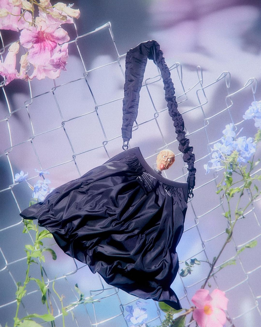 Black shoulder bag with a scrunchie-style strap hanging in front of a chain link fence with colorful floral accents, studio shot.