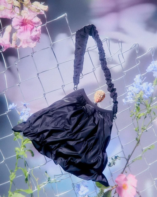 Black shoulder bag with a scrunchie-style strap hanging in front of a chain link fence with colorful floral accents, studio shot.