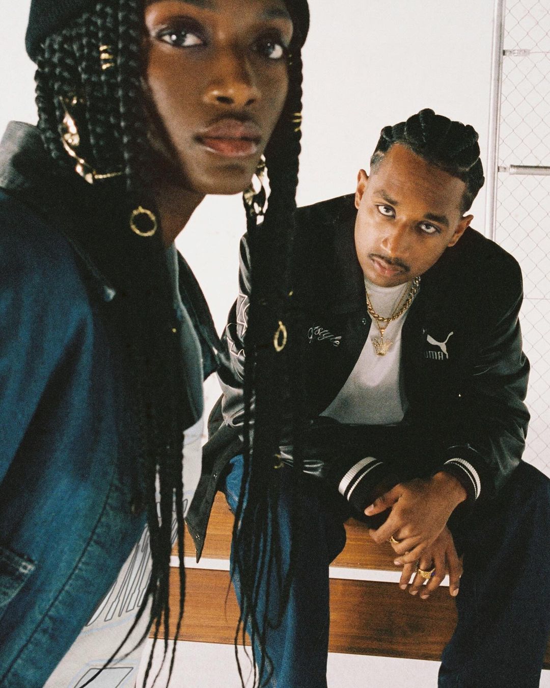 Two stylish young adults pose in front of white wall. One wears a beanie, jacket, and braids with gold rings; the other wears a Puma jacket and gold chain.