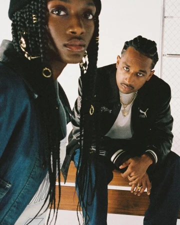 Two stylish young adults pose in front of white wall. One wears a beanie, jacket, and braids with gold rings; the other wears a Puma jacket and gold chain.