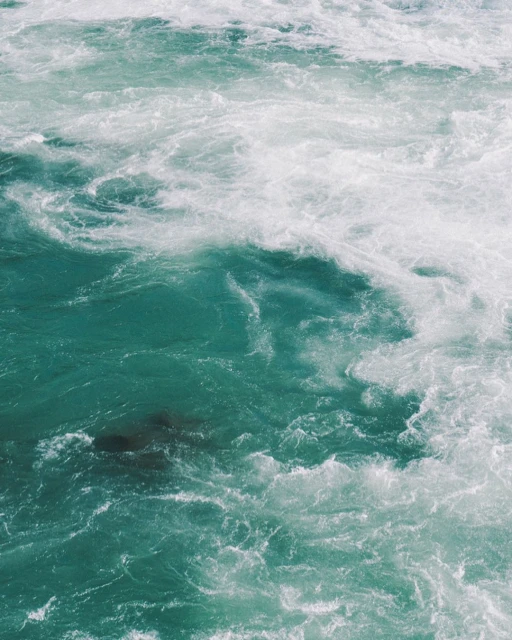Dynamic turquoise water featuring swirling white foam creates an abstract, textured water surface with an energetic, flowing, aquatic feel.