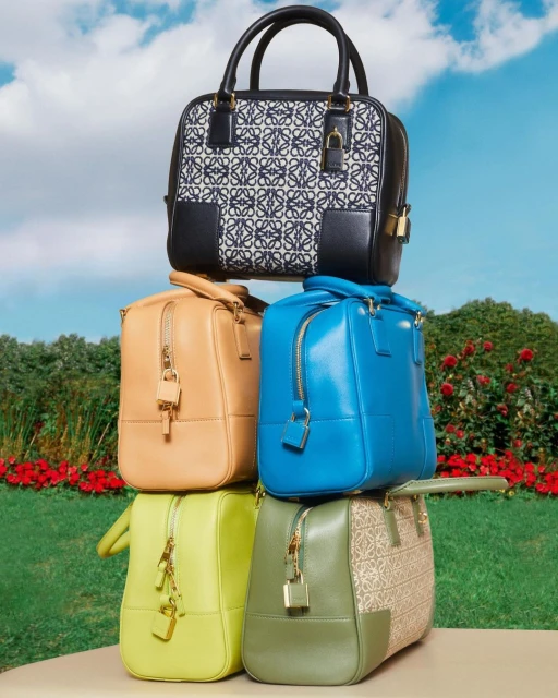 Stack of luxury Loewe handbags in varied colors and designs displayed in a lush outdoor setting with green grass and trees.