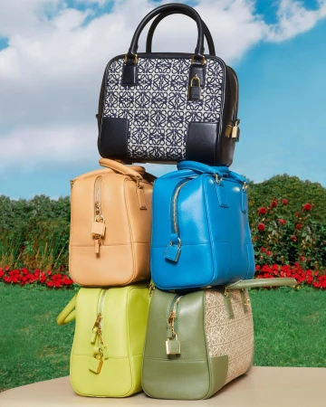 Stack of luxury Loewe handbags in varied colors and designs displayed in a lush outdoor setting with green grass and trees.