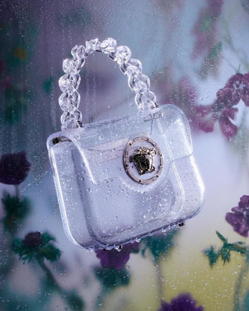 Versace purse: Clear PVC handbag with crystal-like handle and silver Medusa emblem, set against a floral background with water droplets.