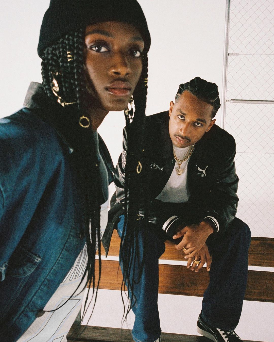 Two models pose indoors against a white wall; the first wears a denim jacket and beanie, the second a Puma jacket and dark jeans.