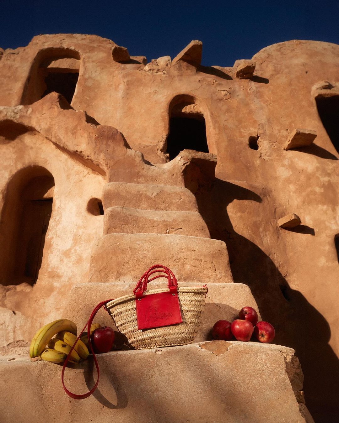 Still life shot of a straw bag with a red panel, bananas, and apples on a staircase against the backdrop of a building with a unique facade.