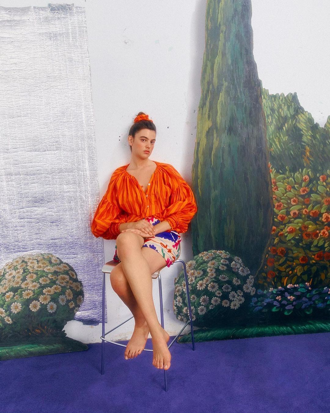 A woman with bare feet sits on a stool wearing an orange top and colorful shorts in front of a painted backdrop inside a studio.