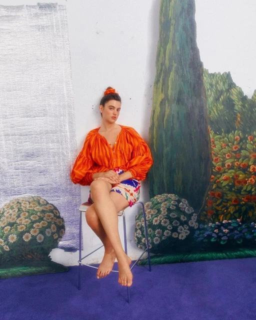 A woman with bare feet sits on a stool wearing an orange top and colorful shorts in front of a painted backdrop inside a studio.