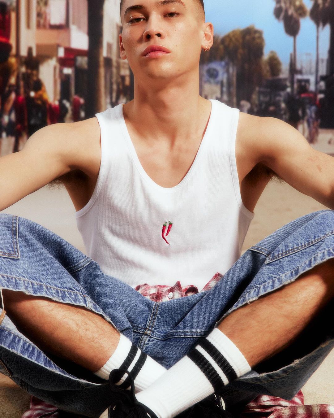 Man in tank top and jeans sitting cross-legged outdoors with palm trees in the background, posing for a fashion shot.
