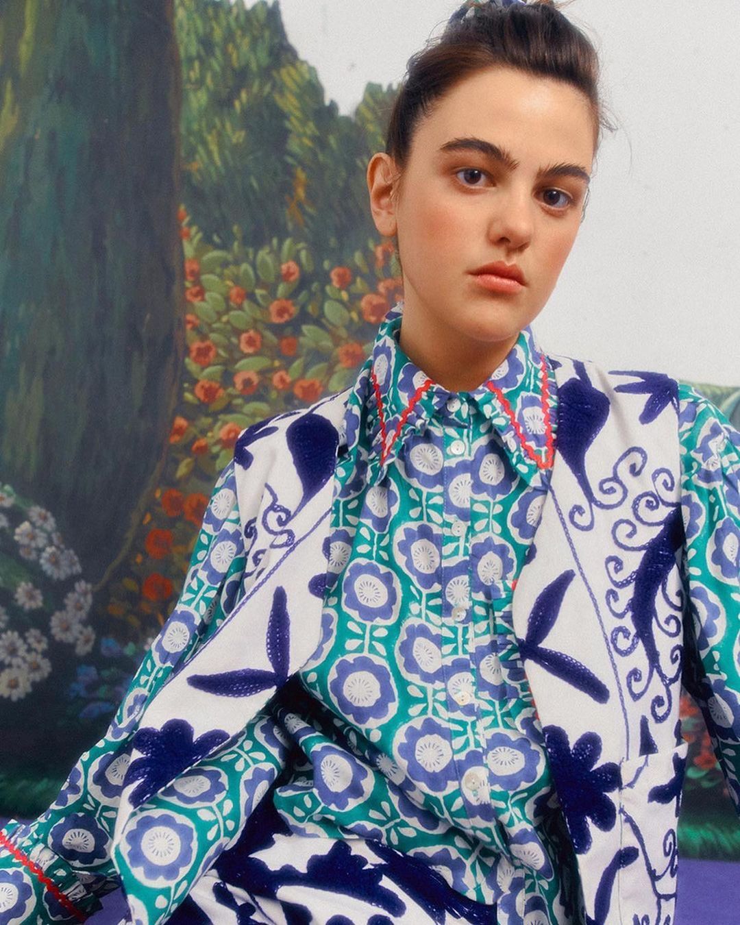 Portrait of a woman wearing a patterned shirt and blazer, posing in front of an artful, colorful backdrop with floral and garden themes.