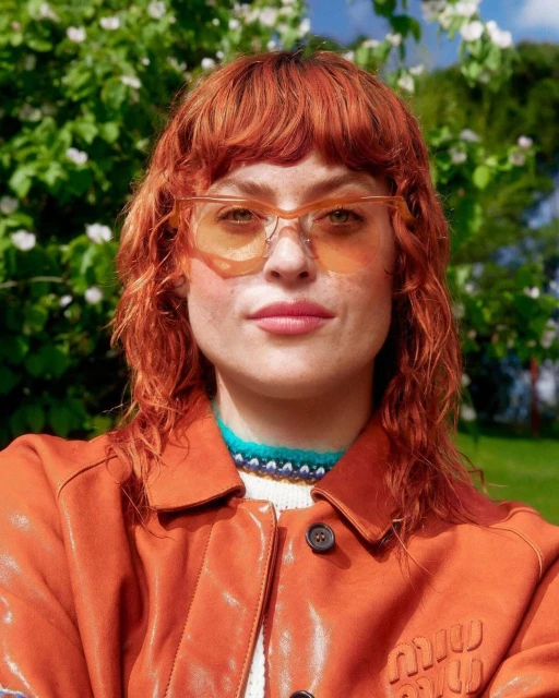 Close-up portrait of a woman with curly red hair, wearing unique translucent glasses and a caramel-colored leather jacket outdoors.