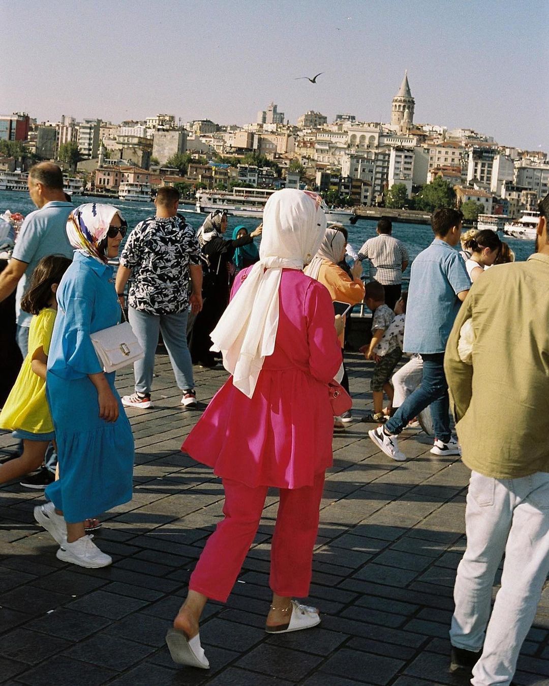 Diverse group of people walk on a sunny day in an urban environment near the water, with buildings visible in the background.