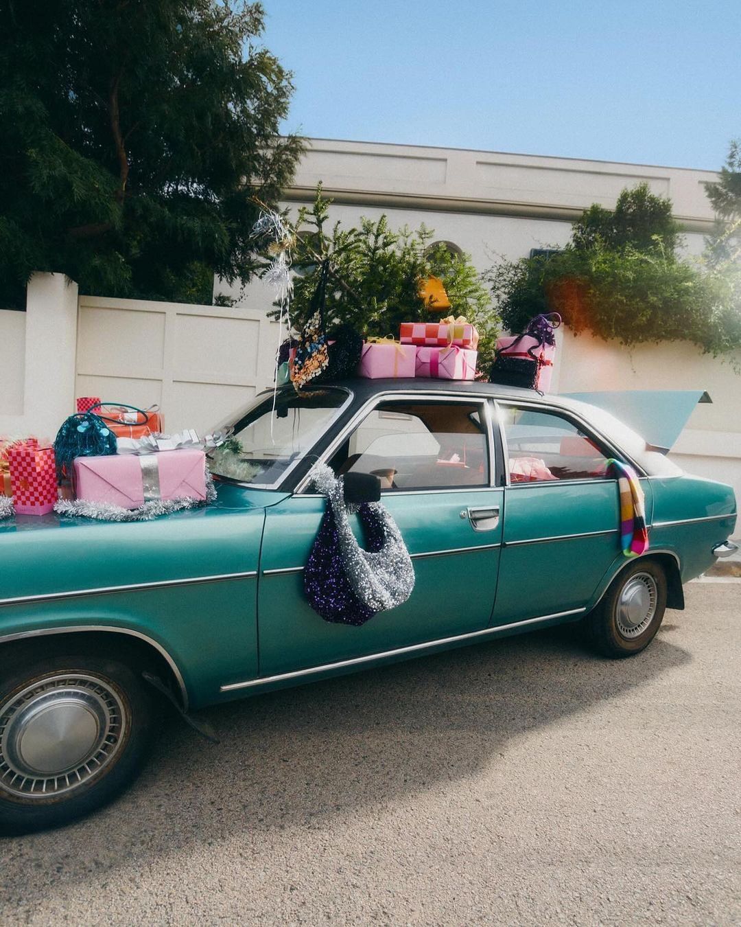 Vintage teal car parked outside a white building, decorated with Christmas presents, greenery, and colorful scarf.