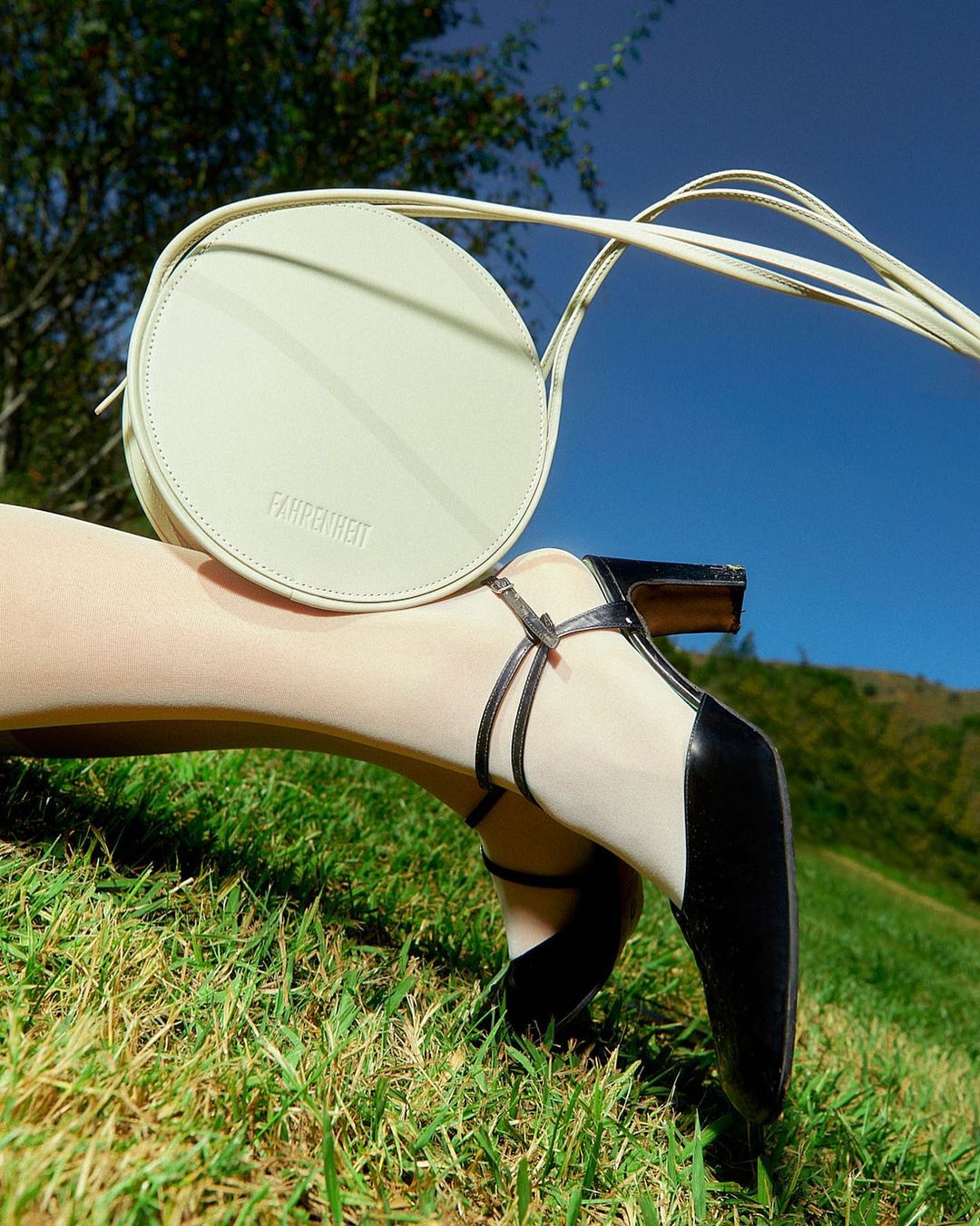 Round handbag on model's leg with heeled shoe on grass with blue sky. Fashion shot on sunny day with bag as primary subject.