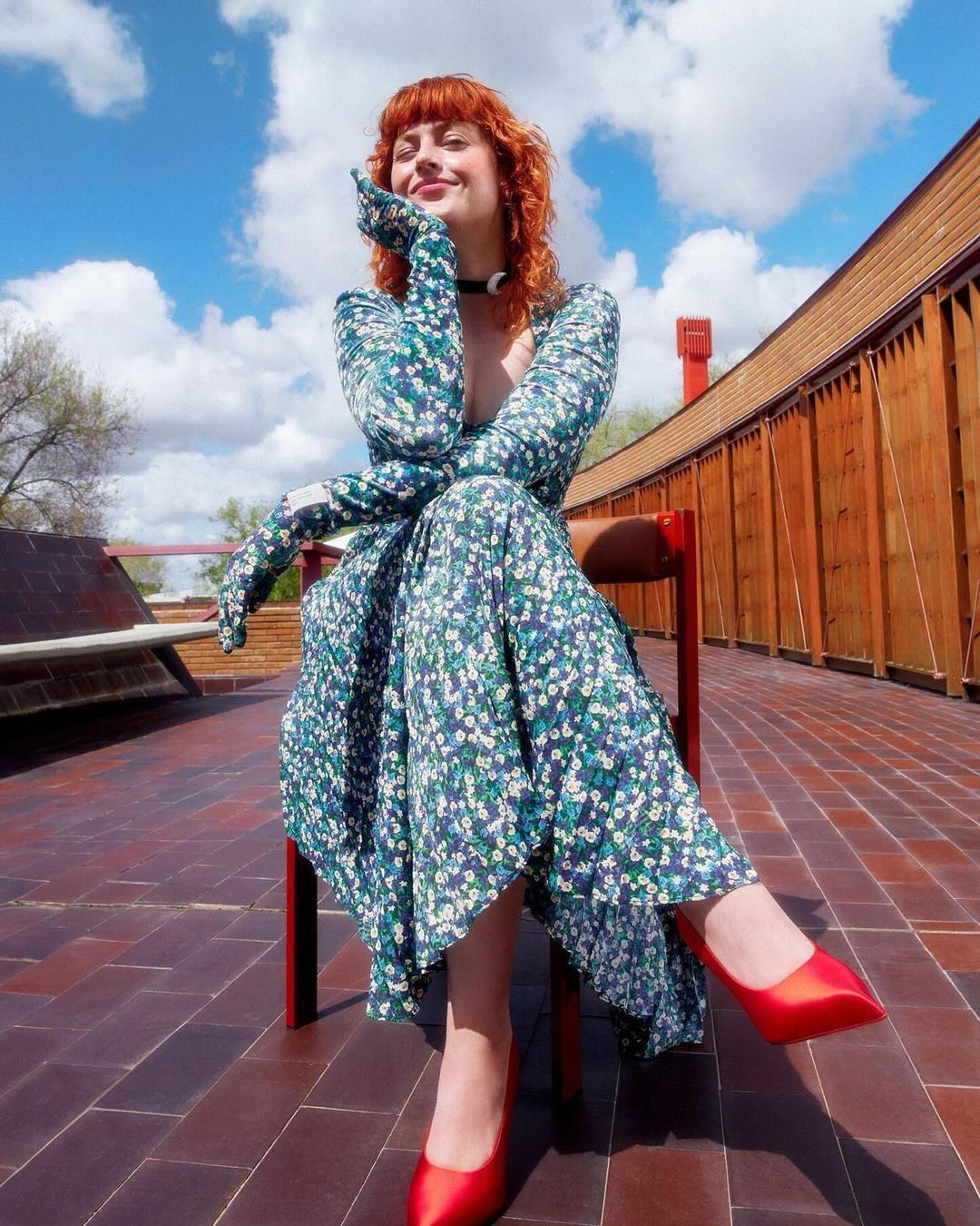 Red-haired woman in a floral dress and red heels poses seated outdoors against a partly cloudy sky with an architectural backdrop.