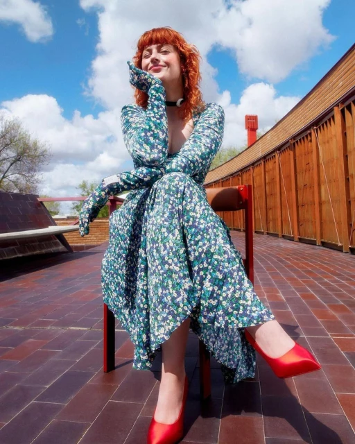 Red-haired woman in a floral dress and red heels poses seated outdoors against a partly cloudy sky with an architectural backdrop.