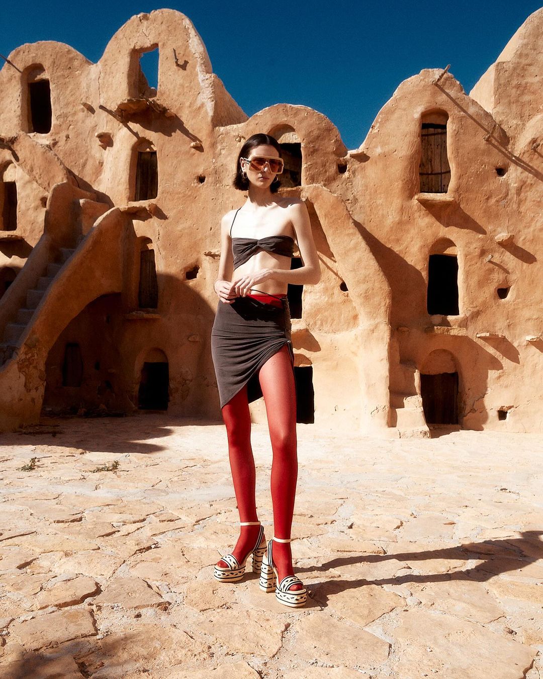 Fashion shot of a woman in a two-piece outfit and platform heels, posing in front of a distinctive earth-toned building under clear skies.