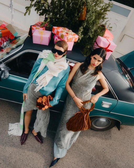 Two women in dresses pose with a Christmas tree and presents on a dark blue car, photographed outdoors in bright daylight.