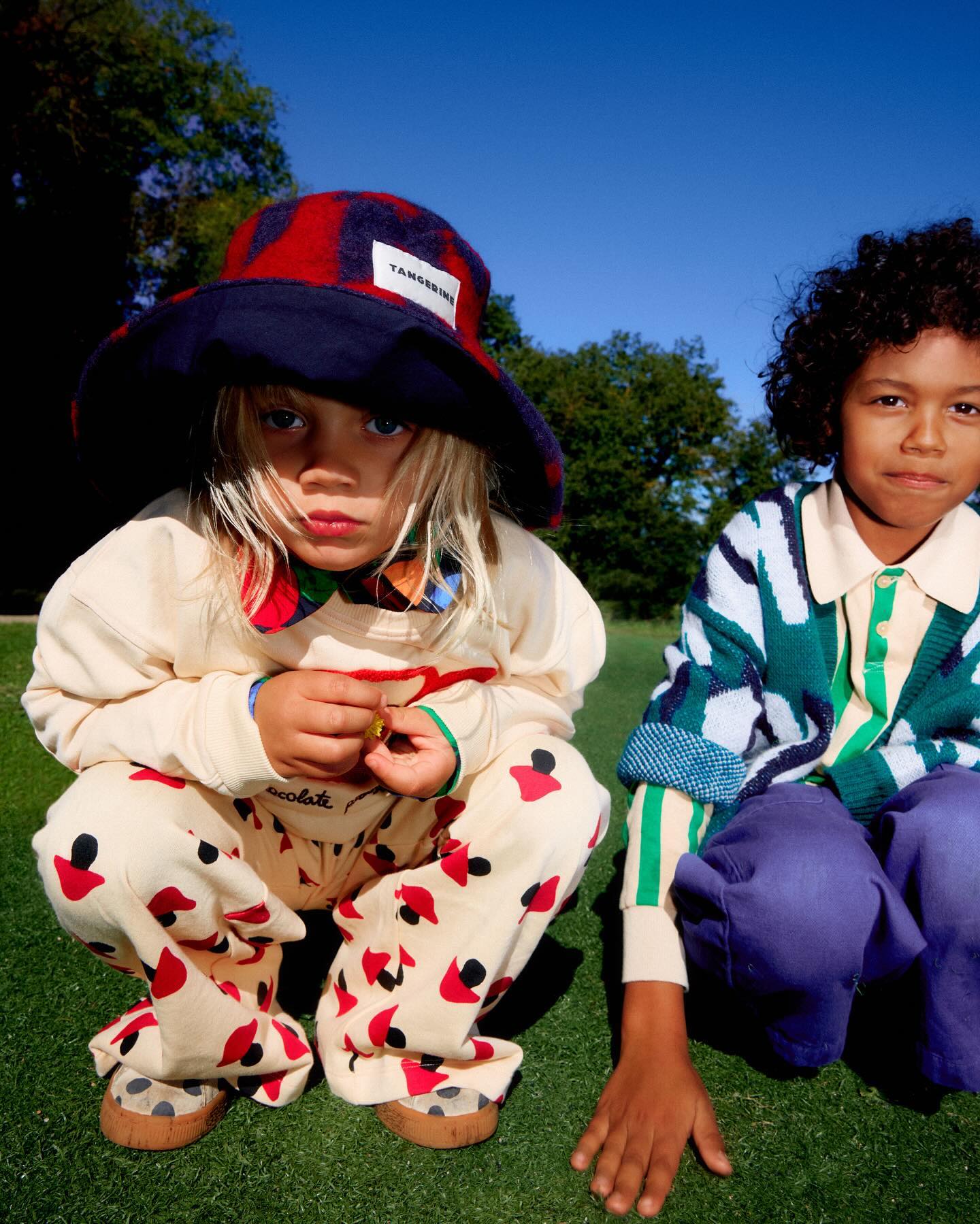 Two children squat on a green lawn, wearing colorful patterned clothing. One child has a bucket hat and a serious expression.