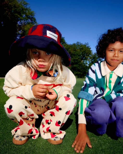 Two children squat on a green lawn, wearing colorful patterned clothing. One child has a bucket hat and a serious expression.