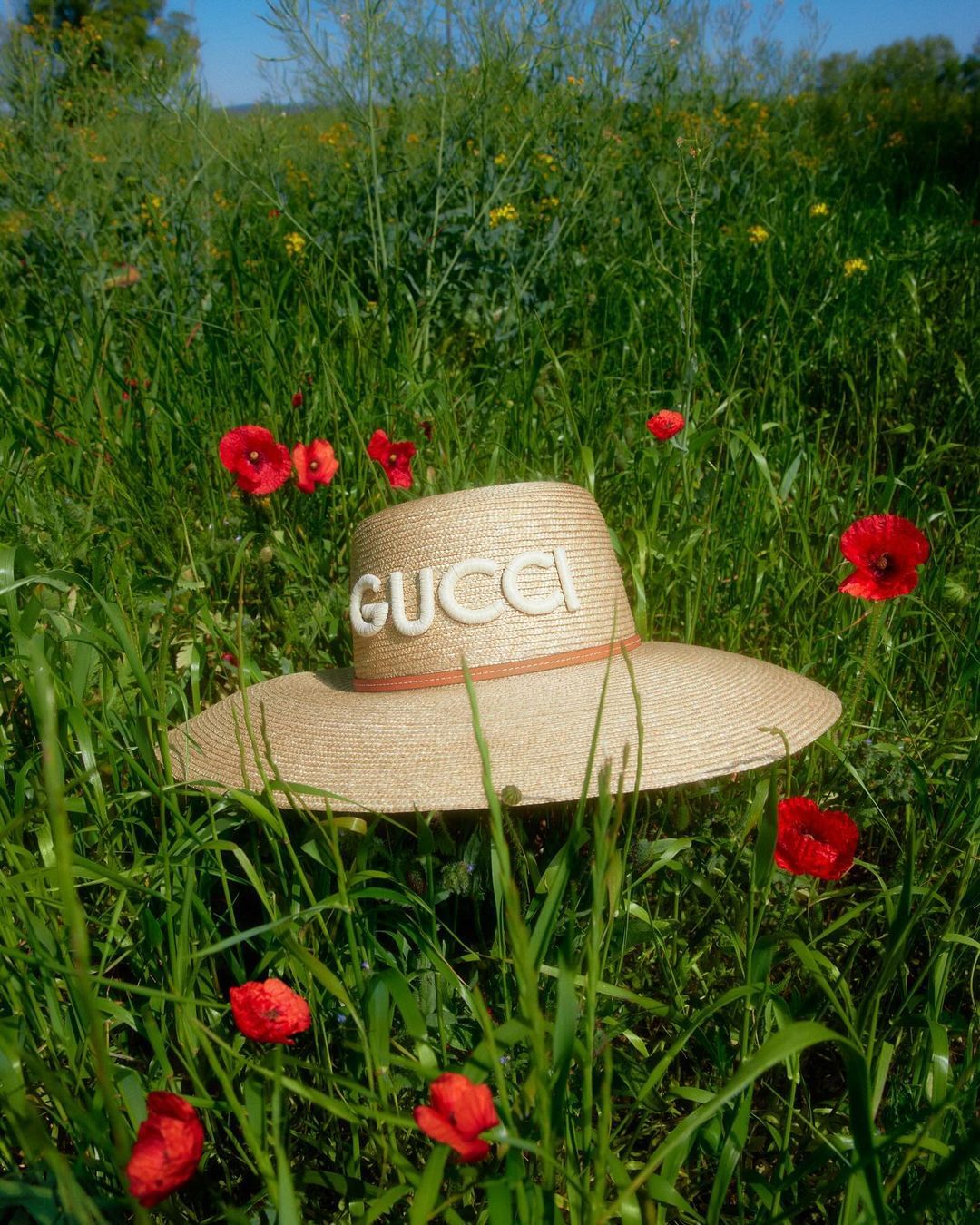 Wide shot of a straw sun hat with the word GUCCI on it, surrounded by tall green grass and red poppy flowers in a sunny field.