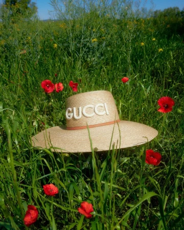 Wide shot of a straw sun hat with the word GUCCI on it, surrounded by tall green grass and red poppy flowers in a sunny field.