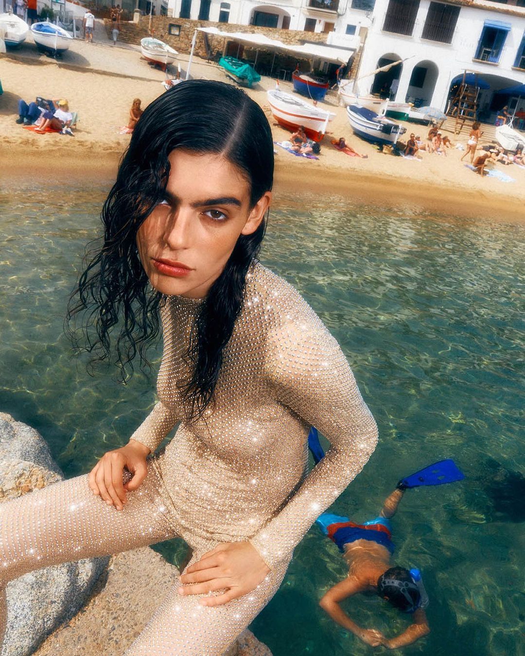 Fashion shot of a woman in a beaded outfit on a rocky shore, with a child snorkeling in the clear water, and boats on the beach.