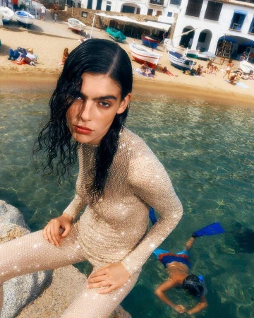 Fashion shot of a woman in a beaded outfit on a rocky shore, with a child snorkeling in the clear water, and boats on the beach.