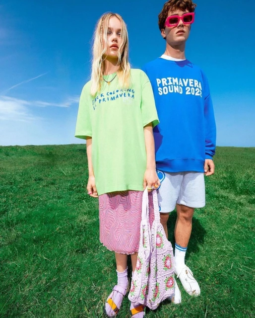 Two young adults stand outside on a sunny day; the woman holds a crocheted bag, and both wear colorful shirts with graphic text.