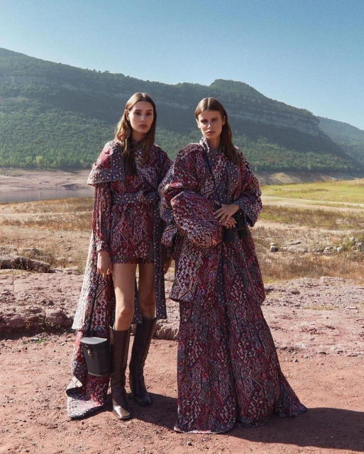 Two women pose in patterned dresses outdoors against a backdrop of mountains and a lake on a sunny day in a fashion editorial.