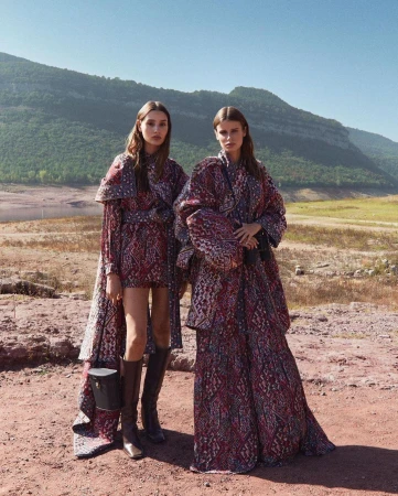 Two women pose in patterned dresses outdoors against a backdrop of mountains and a lake on a sunny day in a fashion editorial.