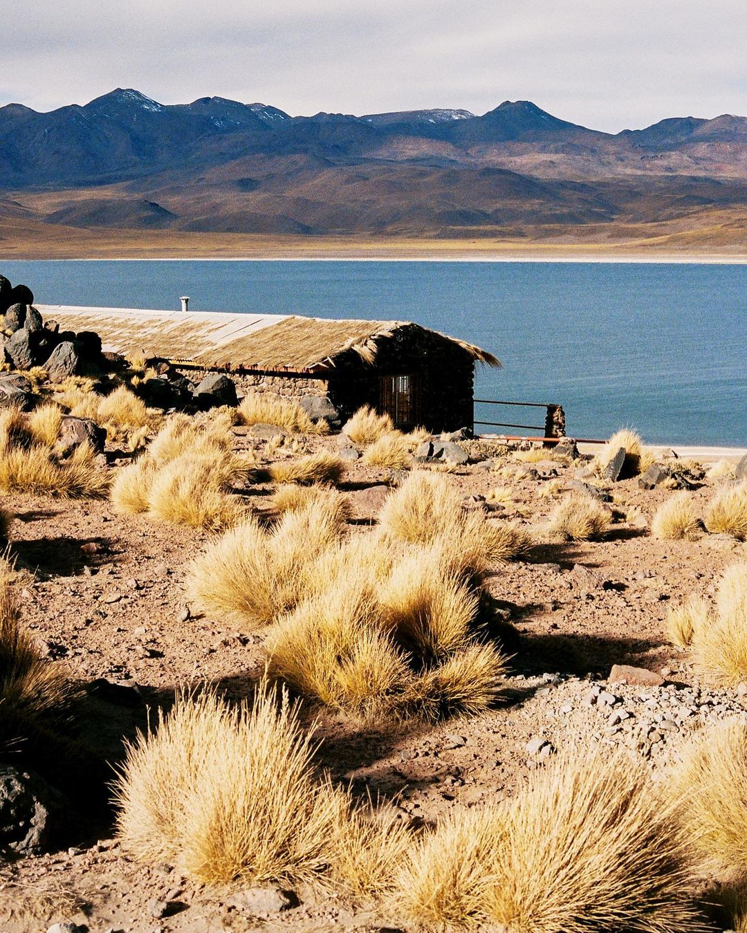 Lakeside cabin nestled in arid landscape, framed by mountains under a bright sky. The structure blends with the natural surroundings.
