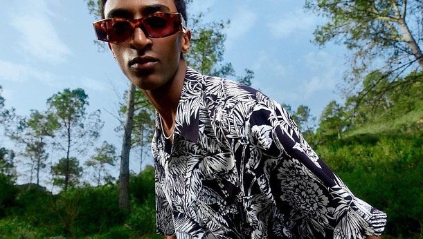 Man wearing a black and white tropical print short-sleeve shirt and red-tinted sunglasses outdoors in a forest.