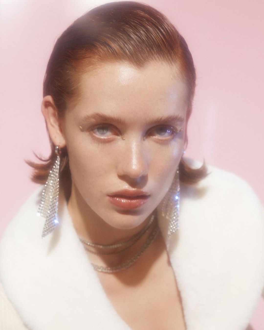 Close-up of an adult female with fair skin, light eyes, and auburn hair, wearing diamond earrings and a white fur coat against a pink background.