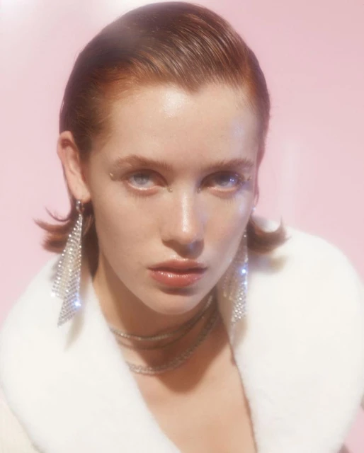 Close-up of an adult female with fair skin, light eyes, and auburn hair, wearing diamond earrings and a white fur coat against a pink background.