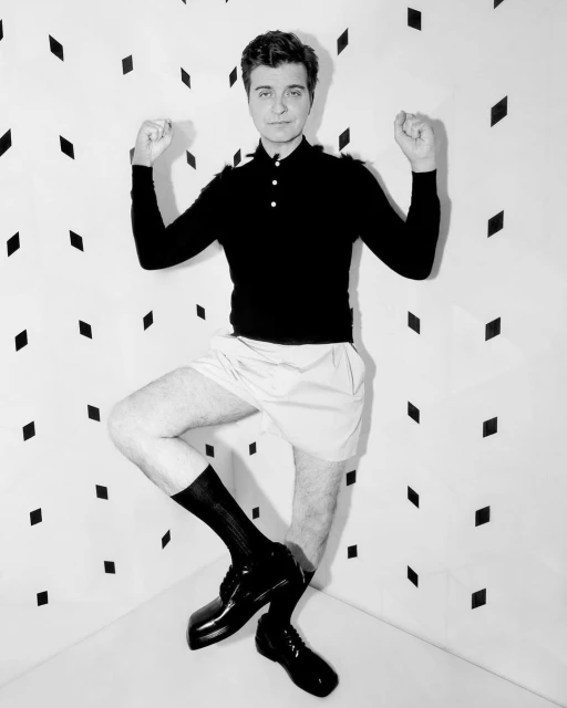 Man poses in photo studio wearing black long sleeve polo shirt, white shorts, black socks, and black shoes against geometric wall backdrop.