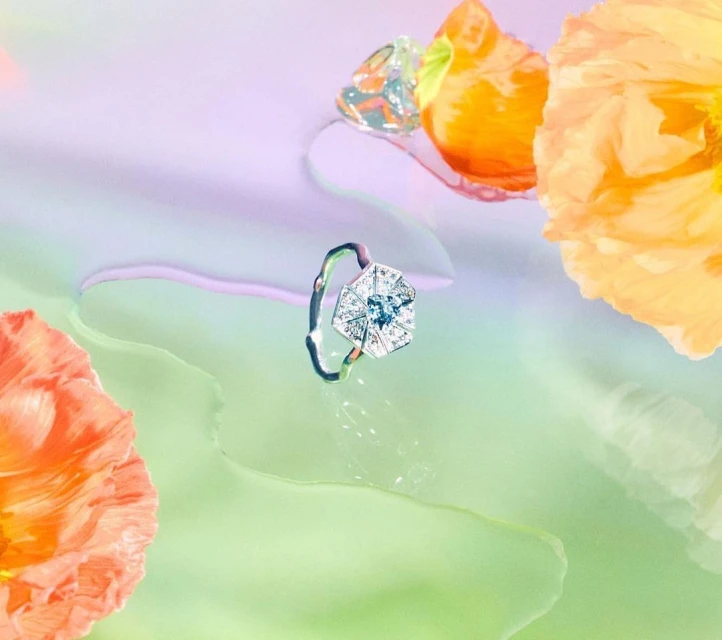 Close-up of a diamond ring surrounded by soft orange flowers against a blend of light green, purple, and white gradient background.