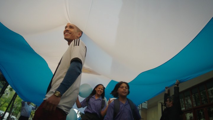 A group including a young man holding up a large Argentinian flag, with two smiling children, captured at eye-level outdoors.