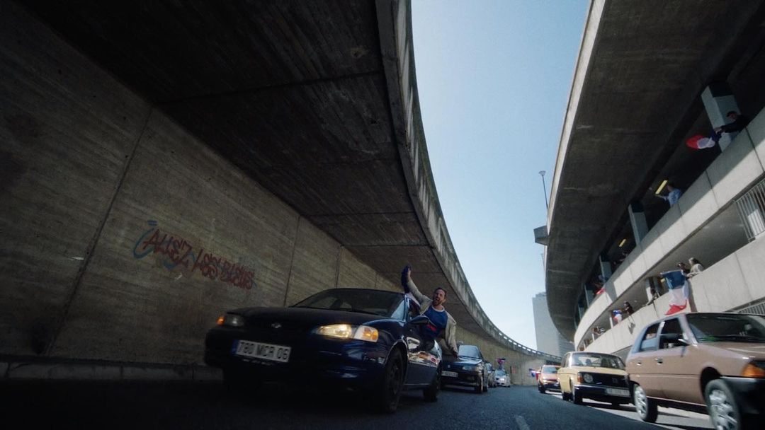 Man perched on a car under an overpass with French flags hanging above, surrounded by cars in an urban setting on a sunny day.