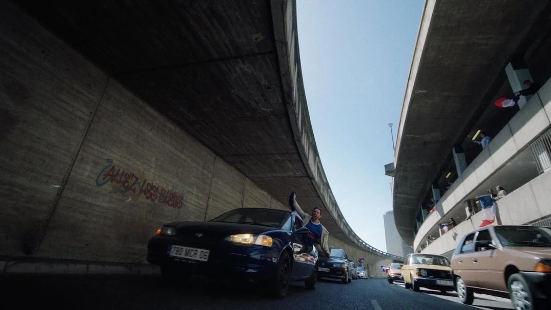 Man perched on a car under an overpass with French flags hanging above, surrounded by cars in an urban setting on a sunny day.
